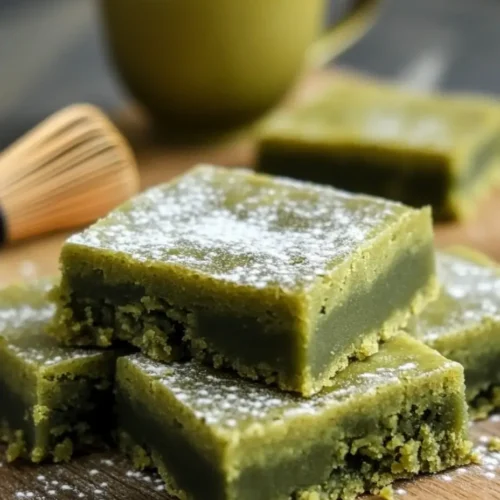 Close-up of matcha mochi brownies dusted with powdered sugar, served on a wooden board with a cup of tea and bamboo whisk in the background.