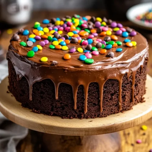 Cosmic brownie-inspired chocolate cake topped with rainbow candy-coated chocolates and drippy ganache on a wooden cake stand.